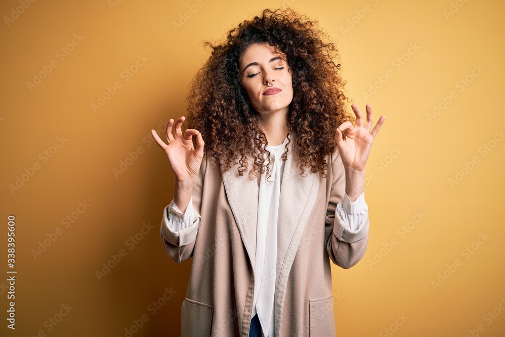 Young beautiful brunette woman with curly hair and piercing wearing casual t-shirt and diadem relax and smiling with eyes closed doing meditation gesture with fingers. Yoga concept. Young beautiful brunette woman with curly hair and piercing wearing casual t-shirt and diadem relax and smiling with eyes closed doing meditation gesture with fingers. Yoga concept.