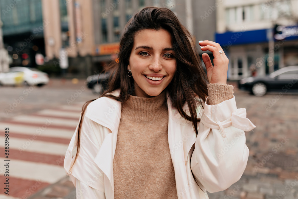 Obraz premium Close up outside portrait of smiling pretty woman with dark hair wearing beige sweater and white coat walking on the street with happy emotions on background of old buildings