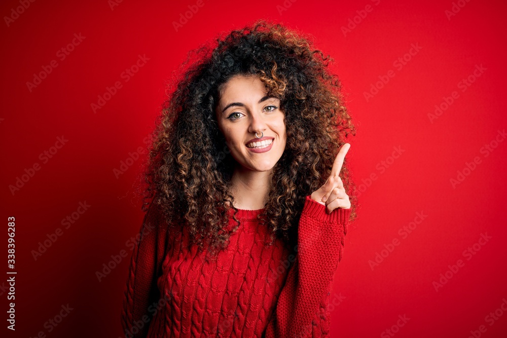 Young beautiful woman with curly hair and piercing wearing casual red sweater showing and pointing up with finger number one while smiling confident and happy. Young beautiful woman with curly hair and piercing wearing casual red sweater showing and pointing up with finger number one while smiling confident and happy.