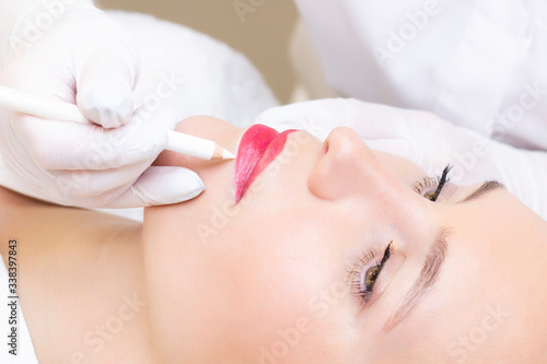 Young woman having permanent makeup on her lips at the beauticians salon. Permanent Makeup (Tattoo). drawing a contour with a white lip pencil