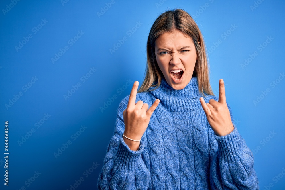 Young beautiful blonde woman wearing winter wool sweater over blue isolated background shouting with crazy expression doing rock symbol with hands up. Music star. Heavy concept. Young beautiful blonde woman wearing winter wool sweater over blue isolated background shouting with crazy expression doing rock symbol with hands up. Music star. Heavy concept.