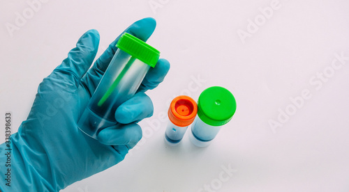 A hand in a blue latex glove holds a container for collecting feces. Plastic container for analysis in the doctors hand. White background.