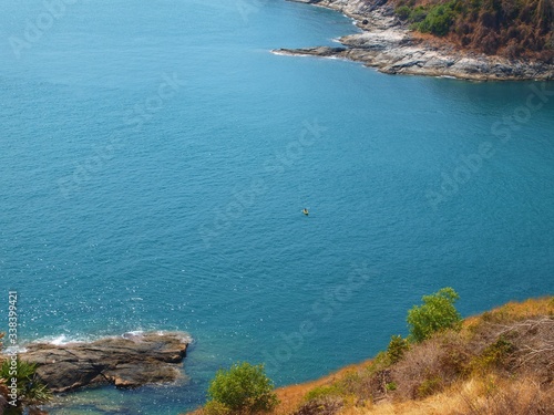 Beautiful view to the sea from a hill, located on the coast. Expanse of azure water and small kayak (canoe) on it. Rocky shore and island, cerulean ocean and paddling canoeist. Turquoise seawater
