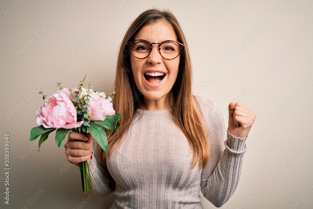 Young blonde woman holding beautiful romantic floral bouquet over isolated background screaming proud and celebrating victory and success very excited, cheering emotion