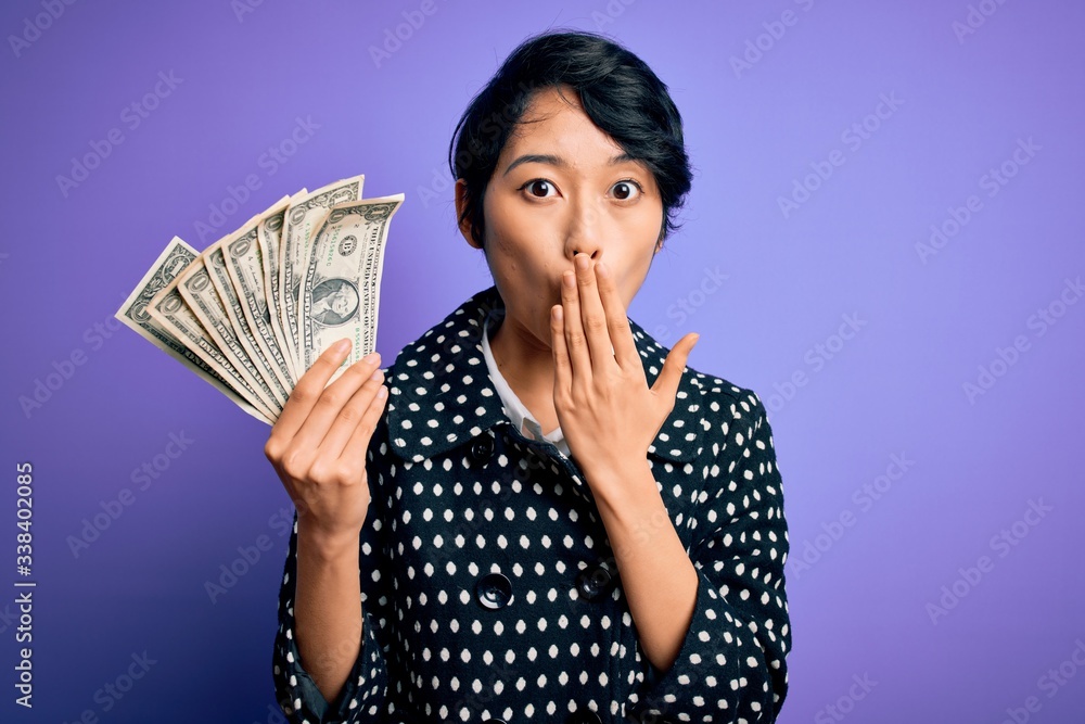Young beautiful asian girl holding bunch of dollars banknotes over purple background cover mouth with hand shocked with shame for mistake, expression of fear, scared in silence, secret concept Young beautiful asian girl holding bunch of dollars banknotes over purple background cover mouth with hand shocked with shame for mistake, expression of fear, scared in silence, secret concept