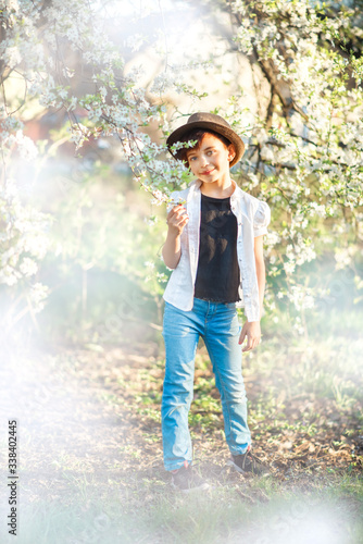 Vertical portrait of 7 years old cute stylish short haired smiling girl in white shirt, blue jeans and brown hat standing in blooming garden in spring. Short haircut for girl. Allergy season © Альбина Саженюк