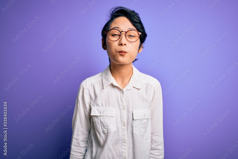 Fototapeta premium Young beautiful asian girl wearing casual shirt and glasses standing over purple background puffing cheeks with funny face. Mouth inflated with air, crazy expression.