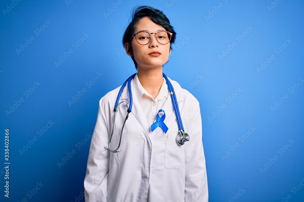 Young beautiful asian doctor girl wearing stethoscope and coat with blue cancer ribbon Relaxed with serious expression on face. Simple and natural looking at the camera. Young beautiful asian doctor girl wearing stethoscope and coat with blue cancer ribbon Relaxed with serious expression on face. Simple and natural looking at the camera.