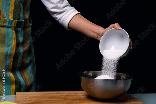 Working in the kitchen. pouring sweet sugar in bowl. Colorful apron. Cooking apple pie. Steel bowl with pieces of fruit. Cook table. Dark black background.