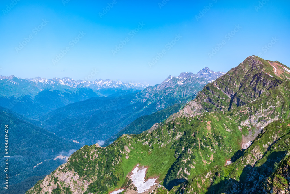 Mountain range with rocks and trees against a clear blue sky. Non-melted snow in the summer on a mountain range. Snow-capped mountain peaks on the horizon
