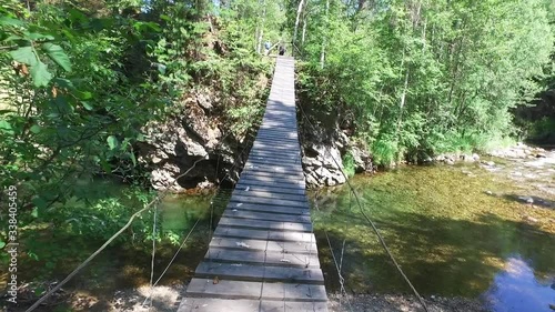 Walking Along Suspension Bridge over the river in Green Forest. Tottering, unsteady bridge. Baikal nature reserve, Russia. Extreme summer Travel. Challenge of hiker scared height. Dangerous adventure.