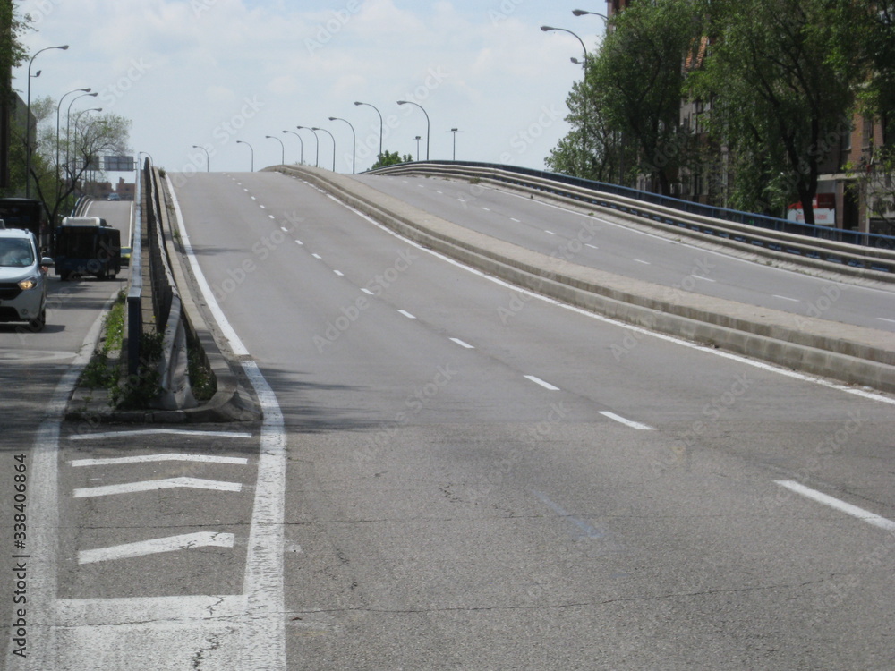 Fototapeta premium Scalextric vacío, vista en perspectiva desde acera,.valla y zona de reserva con separación de vía, señalización en suelo de con líneas blancas, fondo lateral de ciudad con coches ay árboles de ciudad