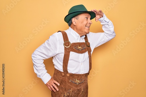 Senior grey-haired man wearing german traditional octoberfest suit over yellow background very happy and smiling looking far away with hand over head. Searching concept.