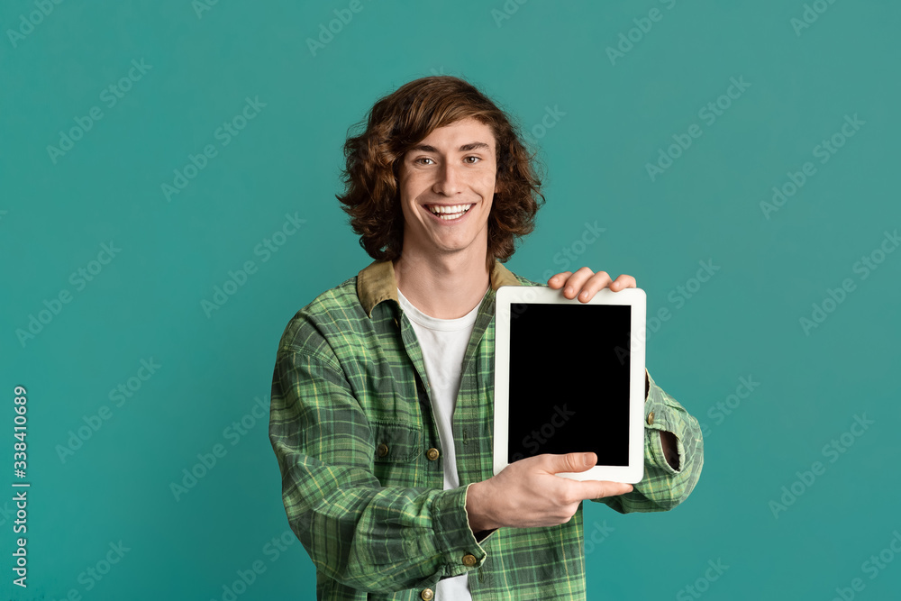Joyful teenage guy showing tablet computer with empty screen on turquoise background, space for design Joyful teenage guy showing tablet computer with empty screen on turquoise background, space for design