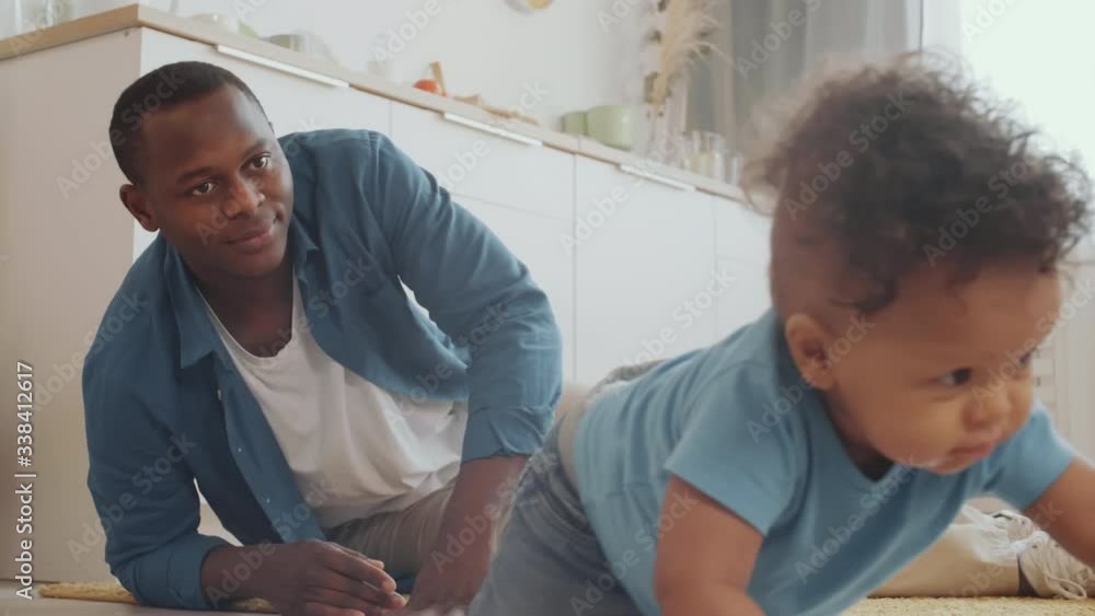 Lockdown of young African man lying on carpet on floor and watching his little curly-haired son creeping Lockdown of young African man lying on carpet on floor and watching his little curly-haired son creeping