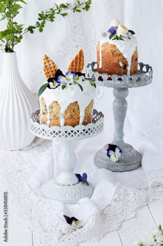 Two Easter cake kulich.Traditional easter sweet bread decorated with hare ears and lilac flowers Viola tricolor on a plate against a white vase with flowering branches. Copy space