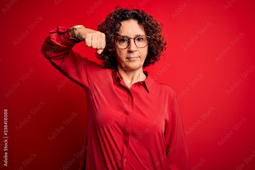 Middle age beautiful curly hair woman wearing casual shirt and glasses over red background Strong person showing arm muscle, confident and proud of power