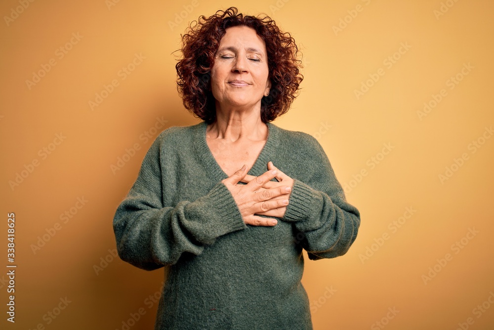 Middle age beautiful curly hair woman wearing casual sweater over isolated yellow background smiling with hands on chest with closed eyes and grateful gesture on face. Health concept. Middle age beautiful curly hair woman wearing casual sweater over isolated yellow background smiling with hands on chest with closed eyes and grateful gesture on face. Health concept.