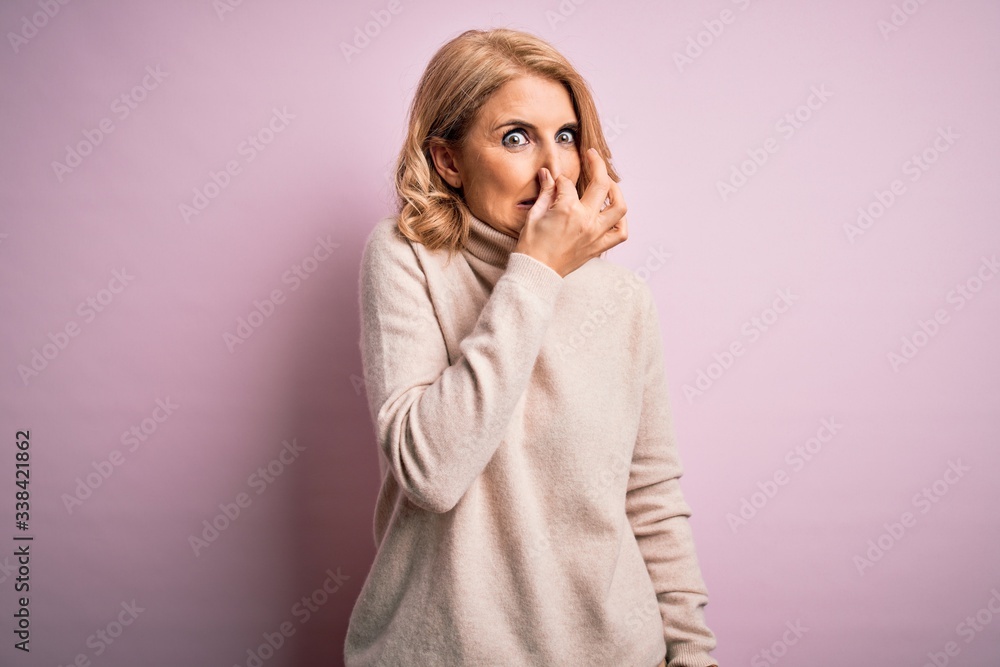Middle age beautiful blonde woman wearing casual turtleneck sweater over pink background smelling something stinky and disgusting, intolerable smell, holding breath with fingers on nose. Bad smell Middle age beautiful blonde woman wearing casual turtleneck sweater over pink background smelling something stinky and disgusting, intolerable smell, holding breath with fingers on nose. Bad smell