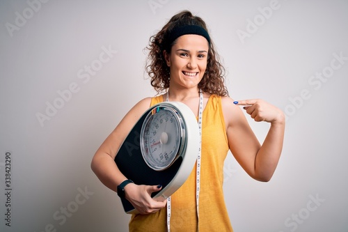 Young beautiful woman with curly hair holding weighing machine and tape measure with surprise face pointing finger to himself
