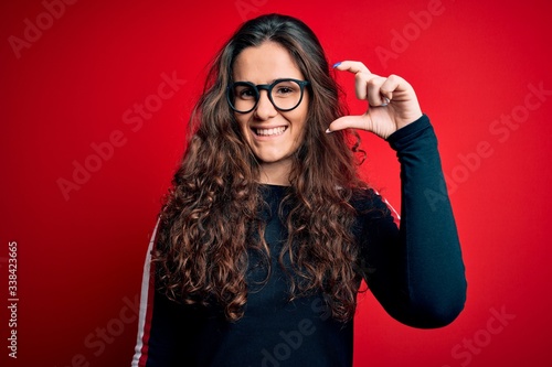 Young beautiful woman with curly hair wearing sweater and glasses over red background smiling and confident gesturing with hand doing small size sign with fingers looking and the camera. Measure