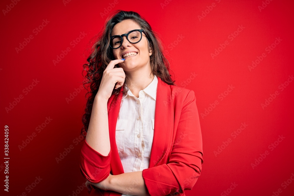 Young beautiful woman with curly hair wearing jacket and glasses over red background looking confident at the camera with smile with crossed arms and hand raised on chin. Thinking positive. Young beautiful woman with curly hair wearing jacket and glasses over red background looking confident at the camera with smile with crossed arms and hand raised on chin. Thinking positive.