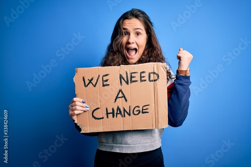Young beautiful activist woman holding banner with change message over blue background screaming proud and celebrating victory and success very excited, cheering emotion