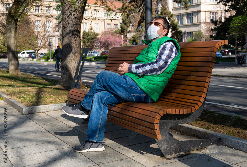 Obraz premium An elderly man in a medical mask sitting on a bench on a deserted embankment during a coronavirus epidemic