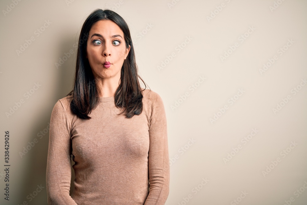 Obraz premium Young brunette woman with blue eyes wearing casual sweater over isolated white background making fish face with lips, crazy and comical gesture. Funny expression.