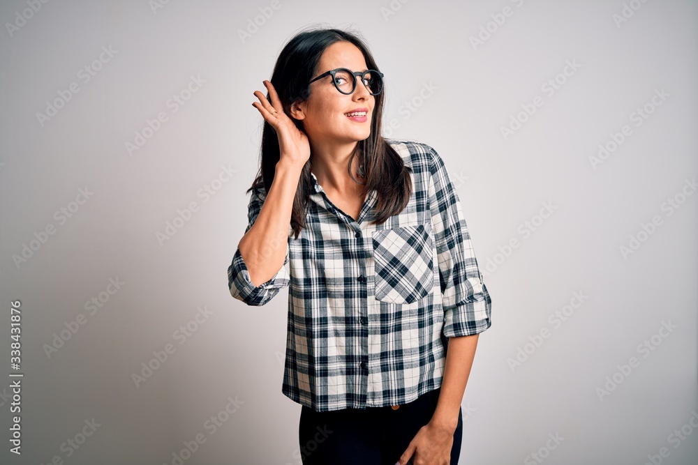 Young brunette woman with blue eyes wearing casual shirt and glasses over white background smiling with hand over ear listening an hearing to rumor or gossip. Deafness concept. Young brunette woman with blue eyes wearing casual shirt and glasses over white background smiling with hand over ear listening an hearing to rumor or gossip. Deafness concept.