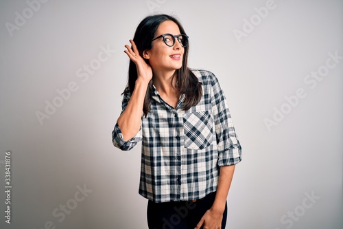 Young brunette woman with blue eyes wearing casual shirt and glasses over white background smiling with hand over ear listening an hearing to rumor or gossip. Deafness concept. © Krakenimages.com