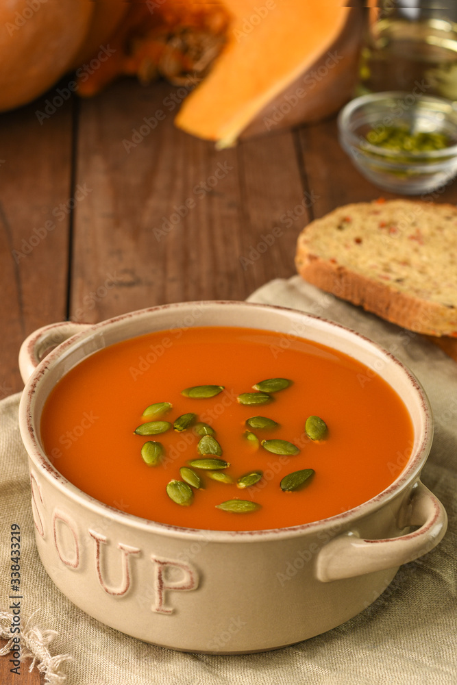 Bowl with pumpkin soup served with seeds on sackcloth with ingredients on wooden background. Carrot organic vegetarian bread, seeds and slice of ripe pumpkin. Menu, recipe Bowl with pumpkin soup served with seeds on sackcloth with ingredients on wooden background. Carrot organic vegetarian bread, seeds and slice of ripe pumpkin. Menu, recipe