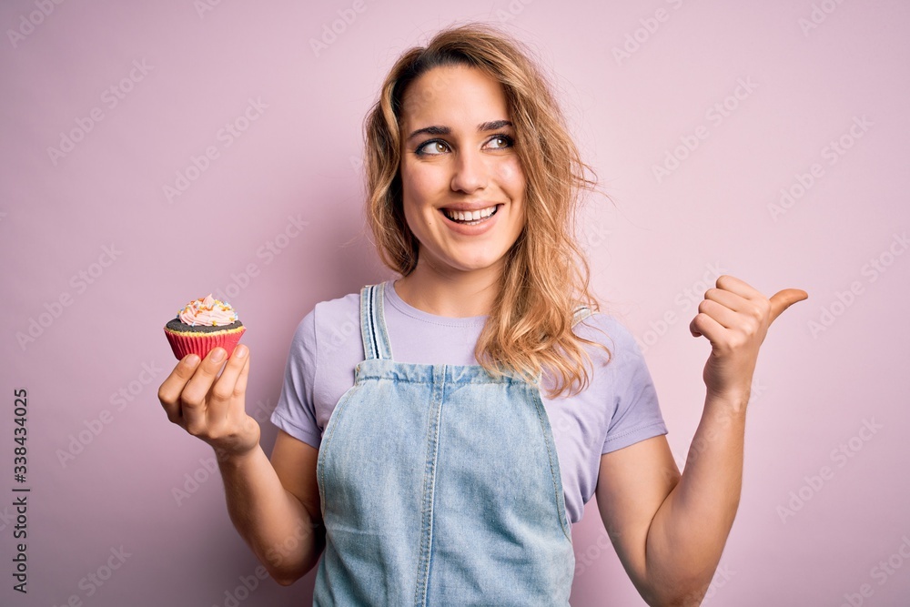 Young beautiful blonde woman eatimg chocolate cupcake over isolated pink background pointing and showing with thumb up to the side with happy face smiling