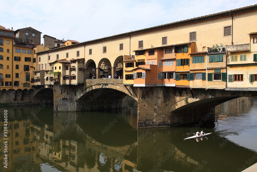 a boat with two rowers runs under a ponte vecchio bridge in florence on the arno river