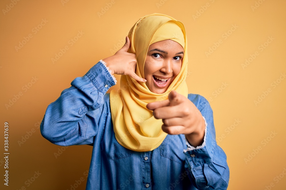 Young beautiful woman with curly hair wearing arab traditional hijab over yellow background smiling doing talking on the telephone gesture and pointing to you. Call me. Young beautiful woman with curly hair wearing arab traditional hijab over yellow background smiling doing talking on the telephone gesture and pointing to you. Call me.