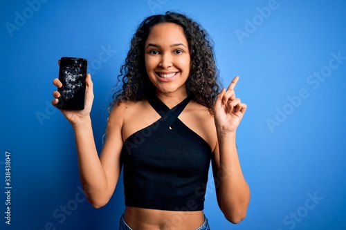 Young beautiful woman with curly hair holding broken smartphone showing cracked screen surprised with an idea or question pointing finger with happy face  number one