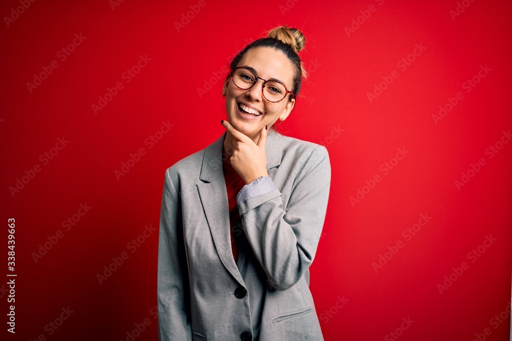 Young beautiful blonde businesswoman with blue eyes wearing glasses and jacket looking confident at the camera smiling with crossed arms and hand raised on chin. Thinking positive.