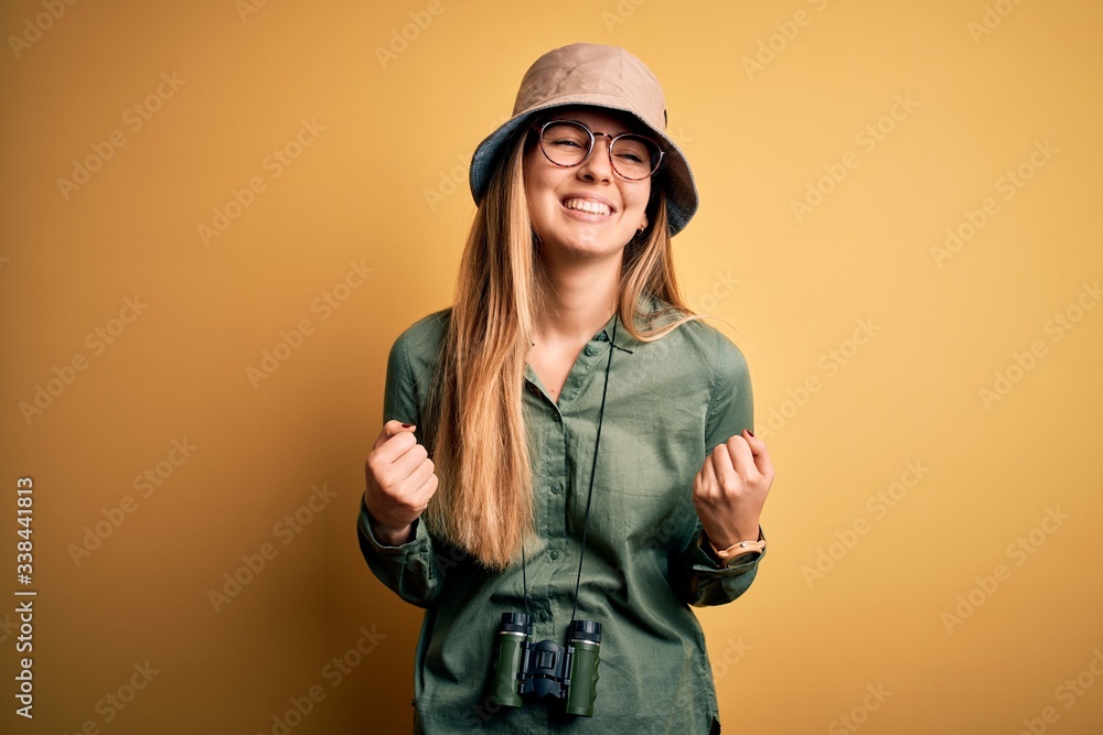 Beautiful blonde explorer woman with blue eyes wearing hat and glasses using binoculars very happy and excited doing winner gesture with arms raised, smiling and screaming for success. Celebration Beautiful blonde explorer woman with blue eyes wearing hat and glasses using binoculars very happy and excited doing winner gesture with arms raised, smiling and screaming for success. Celebration