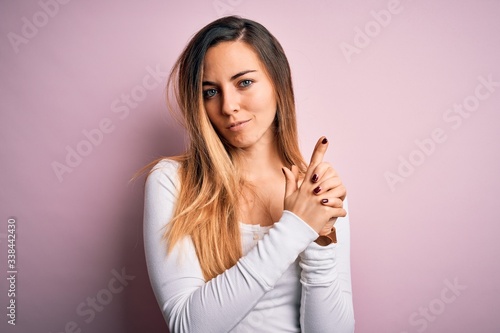 Young beautiful blonde woman with blue eyes wearing white t-shirt over pink background Holding symbolic gun with hand gesture, playing killing shooting weapons, angry face