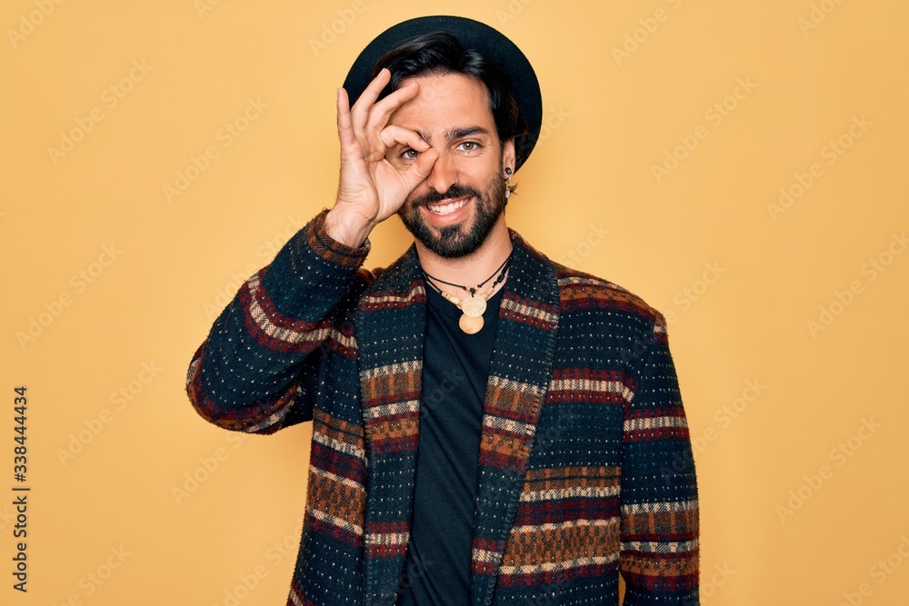 Young handsome hispanic bohemian man wearing hippie style and boho hat doing ok gesture with hand smiling, eye looking through fingers with happy face. Young handsome hispanic bohemian man wearing hippie style and boho hat doing ok gesture with hand smiling, eye looking through fingers with happy face.