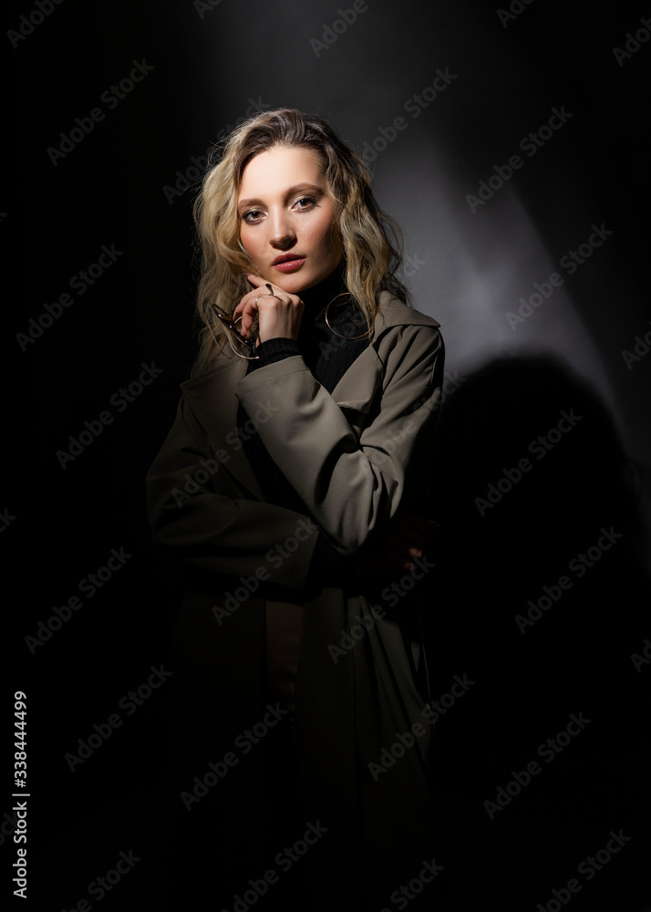 Φωτογραφία Beautiful girl wearing a casual trendy trousers, turtleneck glasses and raincoat posing on a dark background in the shadows