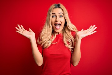 Young beautiful blonde woman wearing casual t-shirt standing over isolated red background celebrating crazy and amazed for success with arms raised and open eyes screaming excited. Winner concept