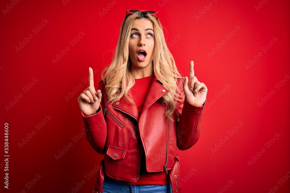 Young beautiful blonde woman wearing casual jacket standing over isolated red background amazed and surprised looking up and pointing with fingers and raised arms. Young beautiful blonde woman wearing casual jacket standing over isolated red background amazed and surprised looking up and pointing with fingers and raised arms.