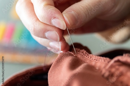 A close up portrait of a hand of someone holding a needle, sewing a hole in a pair of pants at the seam. The needle is about to be put through the fabric to repair it with a thread.