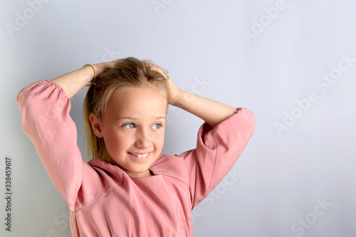 Little blonde girl with long hair in a pink dress posing in front of the camera, makes a ponytail hairstyle, on a gray background, copy space, headshot. Children's fashion and care concept.