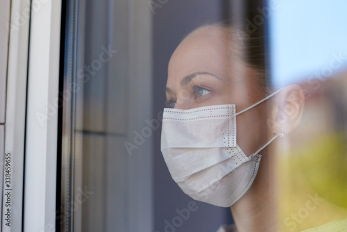 A young sad woman in a medical mask in quarantine looks out the window.