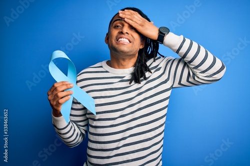 Young african american afro man with dreadlocks holding blue cancer ribbon stressed with hand on head, shocked with shame and surprise face, angry and frustrated. Fear and upset for mistake.