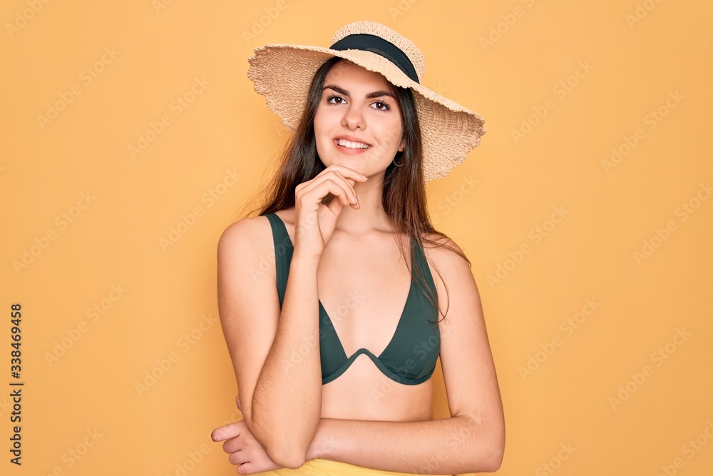 Young beautiful girl wearing swimwear bikini and summer sun hat over yellow background looking confident at the camera with smile with crossed arms and hand raised on chin. Thinking positive. Young beautiful girl wearing swimwear bikini and summer sun hat over yellow background looking confident at the camera with smile with crossed arms and hand raised on chin. Thinking positive.