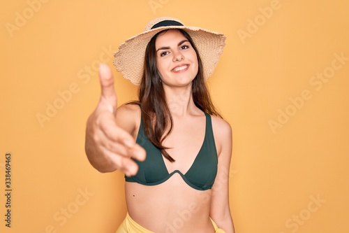 Young beautiful girl wearing swimwear bikini and summer sun hat over yellow background smiling friendly offering handshake as greeting and welcoming. Successful business.