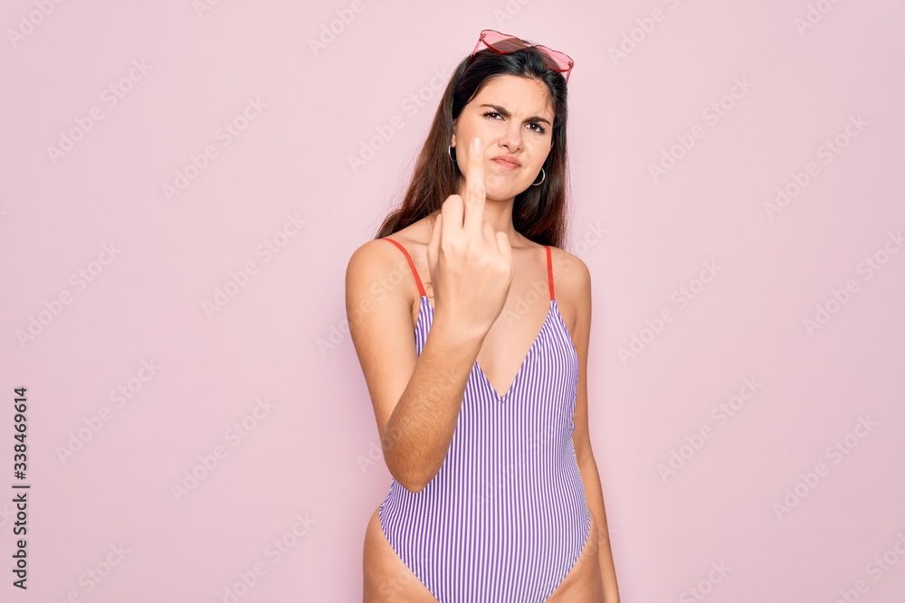 Young beautiful fashion girl wearing swimwear swimsuit and sunglasses over pink background Showing middle finger, impolite and rude fuck off expression Young beautiful fashion girl wearing swimwear swimsuit and sunglasses over pink background Showing middle finger, impolite and rude fuck off expression